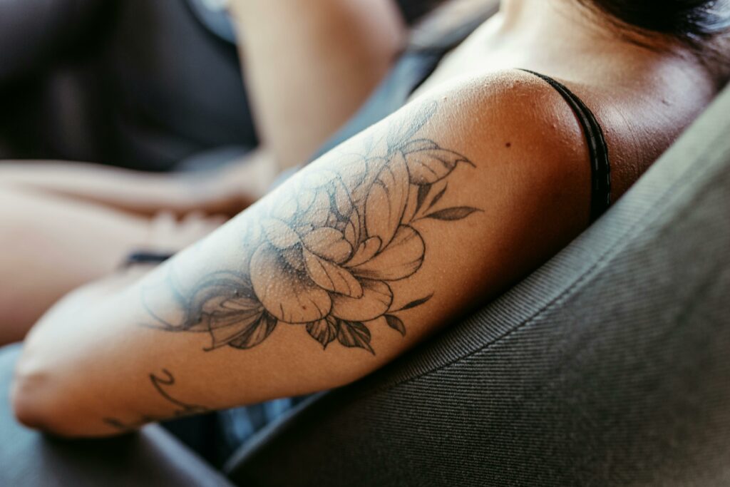 Close-up of a woman's arm with a detailed floral tattoo while she sits relaxed in a chair.
