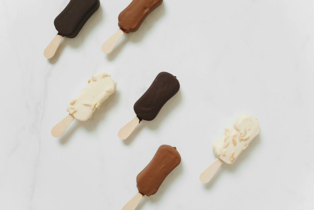 Top view of assorted chocolate and vanilla ice cream bars on wooden sticks placed on a white surface.